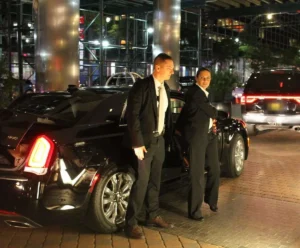 Two close protection officers waiting by a vehicle at a hotel entrance at night
